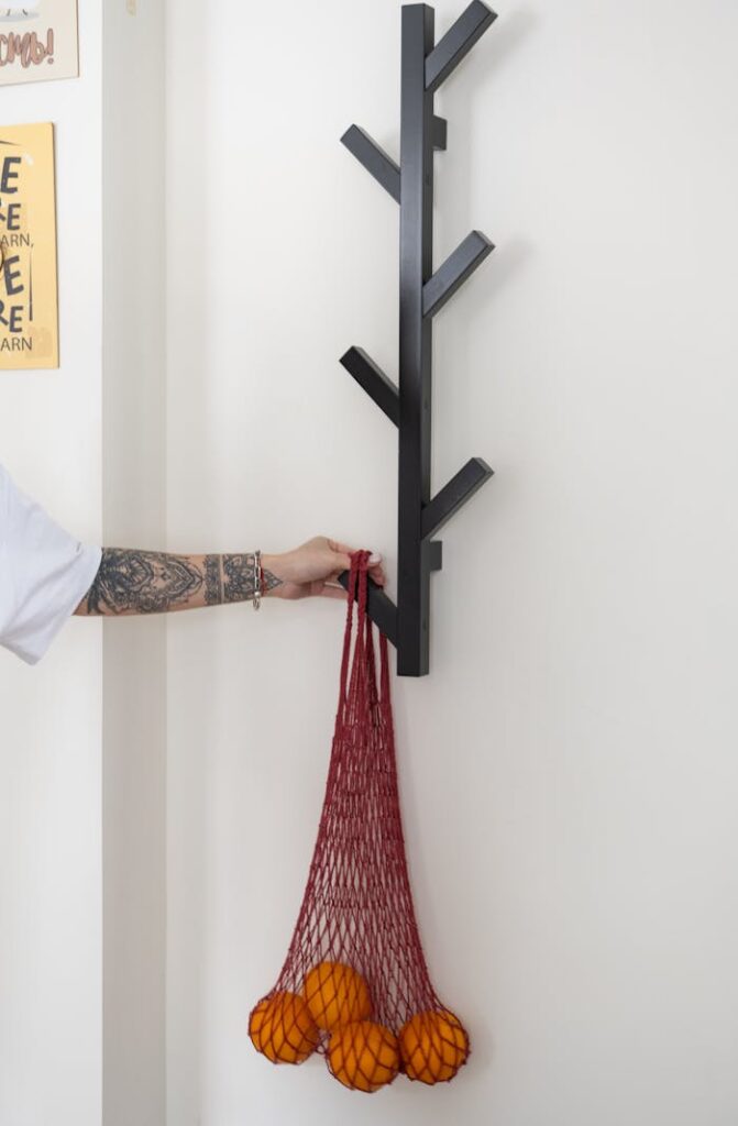 A tattooed hand holds a red net bag filled with oranges, hanging on a modern wall hook.